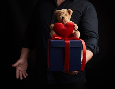 adult man in a black shirt holds a blue square box tied with a red ribbon and sits on top of a brown teddy bear with a heart, concept of congratulations on Valentine's Day on February 14