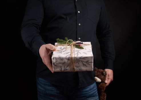 adult man in a black shirt holds a square box and brown teddy bear, concept of congratulations on Valentine's Day on February 14, low key 