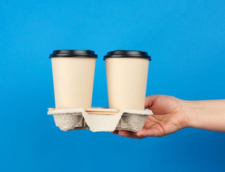 Two brown paper disposable cups with a plastic lid stand in the tray, female hand holds disposable tableware on a blue background