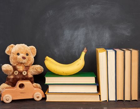 brown teddy bear sits on a brown wooden table and wooden car, behind an empty black chalk board, place for an inscription, back to school