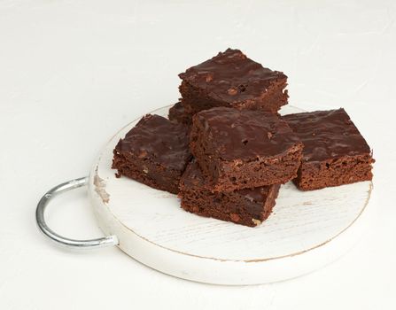 stack of square baked slices of brownie chocolate cake with walnuts on a round white wooden board, top view