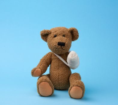 funny vintage brown curly teddy bear with rewound paw with white gauze bandage sitting on blue background, concept of injuries in children or animals