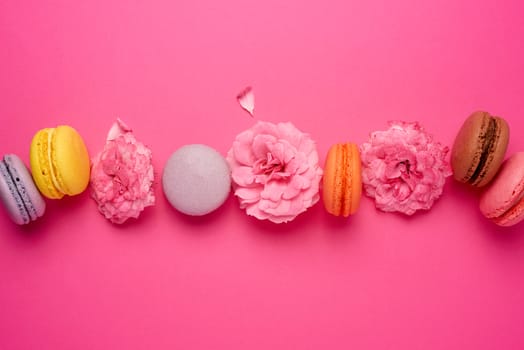 sweet multi-colored macarons with cream and a pink rose bud on a pink background, top view, flat lay