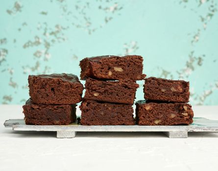 stack of square baked pieces of brownie chocolate cake with walnuts on a wooden board, green background