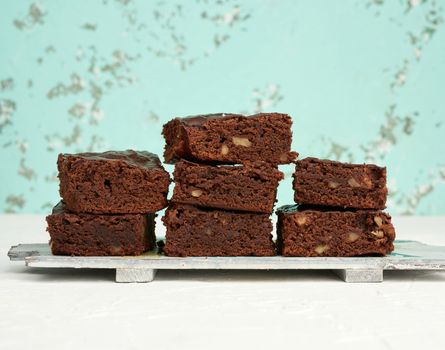 stack of square baked pieces of brownie chocolate cake with walnuts on a wooden board, green background