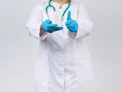 woman physician in a white coat and mask holding a white plastic jar for the medicines and tablets on hand wearing blue latex gloves, white background