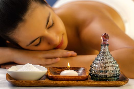 Close up still life of massage oil, candle and Himalayan salt. Out of focus woman relaxing with eyes closed in background.