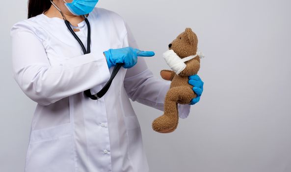 woman in white coat with buttons holding a brown teddy bear with a white gauze bandage tied with her paw, concept of pediatrics and animal treatment