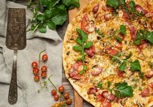 baked round pizza with smoked sausages, mushrooms, tomatoes, cheese and arugula leaves, food is cut in portions, wooden table 