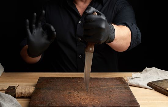 chef in a black shirt and black latex gloves holds a large kitchen knife for cutting meat, right hand shows ok symbol, low key