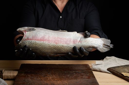 chef in a black shirt and black latex gloves holds a raw carcass of headless salmon fish over a brown wooden cutting board, process of cutting fish