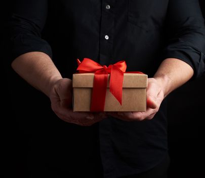 adult man in a black shirt holds in his hand a square brown boxlet tied with a red ribbon on a black background, concept of holiday greetings, surprise and gratitude