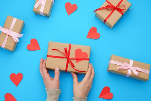 female hand holds a box wrapped in brown kraft paper and tied with a silk ribbon on a blue background, paper red hearts. Valentine's Day backdrop