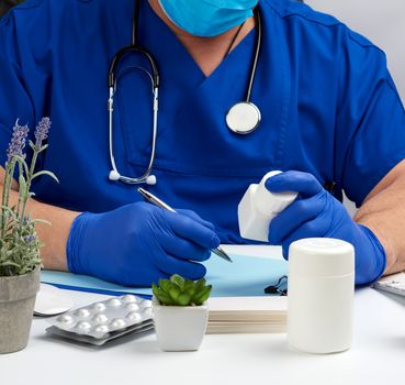 male doctor in a blue uniform sits at a white table and writes in a paper notebook, wearing sterile gloves on his hands, concept of receiving patients in the office