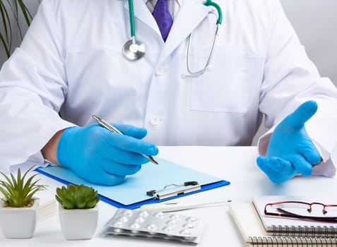 doctor in a white uniform sits at a white table and writes in a paper notebook, wearing sterile gloves on his hands, concept of receiving patients in the office