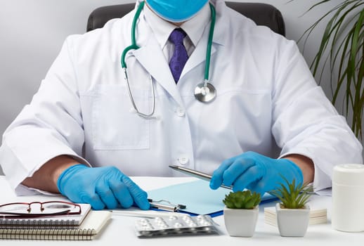doctor in a white uniform sits at a white table and writes in a paper notebook, wearing sterile gloves on his hands, concept of receiving patients in the office