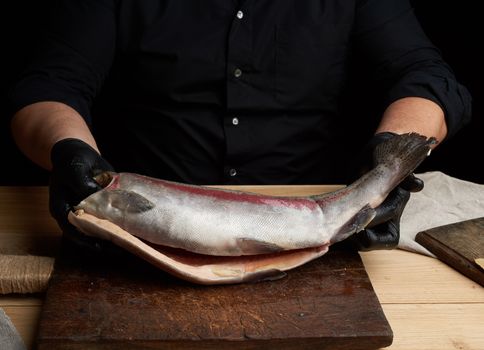 chef in a black shirt and black latex gloves holds a raw carcass of headless salmon fish over a brown wooden cutting board