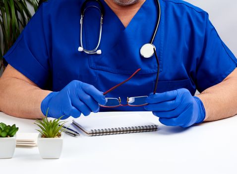 male doctor in blue uniform and blue latex gloves sits at a white work table in his office, concept of patient reception in the clinic, therapist's workplace