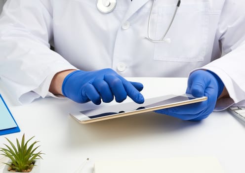 doctor in a white medical coat sits at a table and holds an electronic tablet in his hand, touches the screen with his right hand, doctor’s office, distance medicine