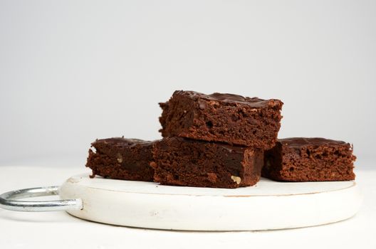 stack of square baked slices of brownie chocolate cake with walnuts on a round white wooden board, close up