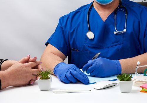 male doctor in uniform and in blue latex medical gloves sits at the table and carries out the reception of patients, workplace and office of the doctor