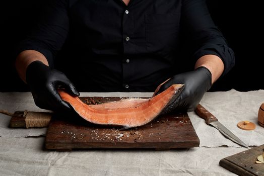 chef in a black shirt and black latex gloves holds a large piece of salmon fish fillet over the table, cooking process, clouse up