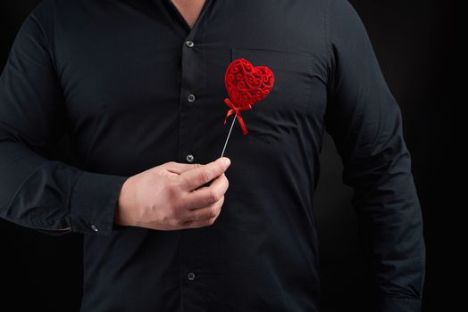 adult man stands on a dark background wearing a black shirt and holding a red carved heart near his chest in his right hand, concept of love