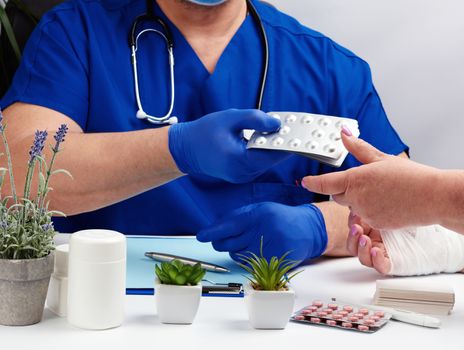 doctor in uniform and blue latex medical gloves sits at a table and examines a patient with a hand injury, doctor gives painkillers, office for patients