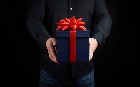 adult man in a black shirt holds a blue square box with a red bow tied on a dark background, concept of congratulations, surprise