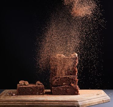 stack of square baked brownie pieces sprinkled with cocoa powder, particles froze in the air against a dark background,  delicious chocolate dessert