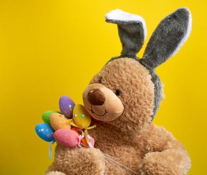 big cute brown teddy bear holding colorful Easter eggs, wearing a rabbit mask with long ears on his head, funny holiday card, close up