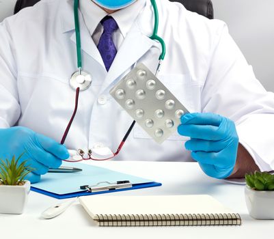 male hand with blue sterile gloves holds a stack of pills in a blister, concept for drug treatment of diseases, doctor sits in an armchair at a desk, office