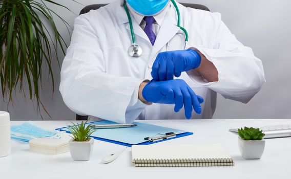 doctor in a white medical coat sits at a table in a brown leather chair and puts on sterile medical gloves in his hands before receiving and examining patients