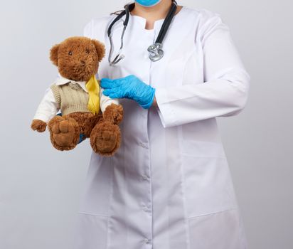 pediatrician in white coat, blue latex gloves holds a brown teddy bear with a yellow ribbon on a sweater, concept of the fight against childhood cancer, problem of suicides