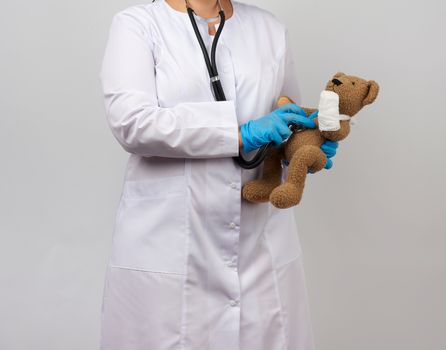 female medic holds brown teddy bear with paw bandaged in white bandage and listens to toy with stethoscope, concept of pediatrics and animal treatment