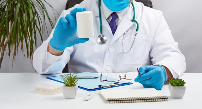 male doctor in a white coat and blue latex gloves sits at a white desk in his office and holds a plastic white jar of pills, concept for receiving patients from a doctor