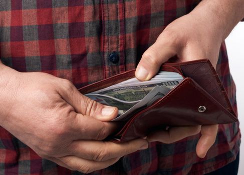 man in a checkered red shirt holds a brown leather wallet full of one hundred US paper dollars banknotes, concept of salary, wealth