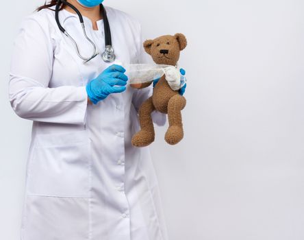 female medic in a white coat with buttons holding a brown teddy bear and bandaging her paw with a white gauze bandage, concept of pediatrics and animal treatment