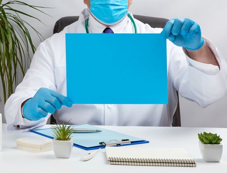 doctor in a white medical coat is sitting at a table in a brown leather chair and holding a empty blue sheet of paper in his hands, place for an inscription, medical office