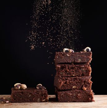 stack of square baked brownie pieces sprinkled with cocoa powder, particles froze in the air against a dark background,  delicious and delicious chocolate dessert
