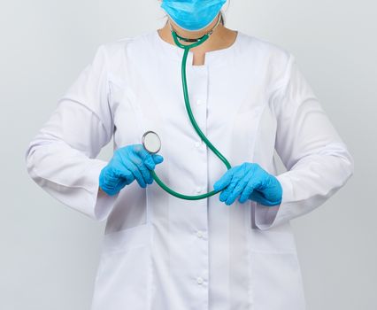 medic woman in a white coat and mask, wearing blue medical latex gloves, holds a green stethoscope on a white background, studio photo
