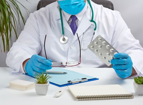 male doctor in a white coat and blue latex gloves sits at a white desk in his office and shows a pack of pills in a blister, concept for drug treatment of diseases, doctor sits in an armchair at a desk