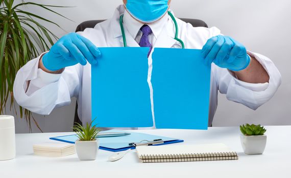 doctor in a white medical coat is sitting at a table in a brown leather chair and holding a torn blue sheet of paper in his hands, place for an inscription, a medical office