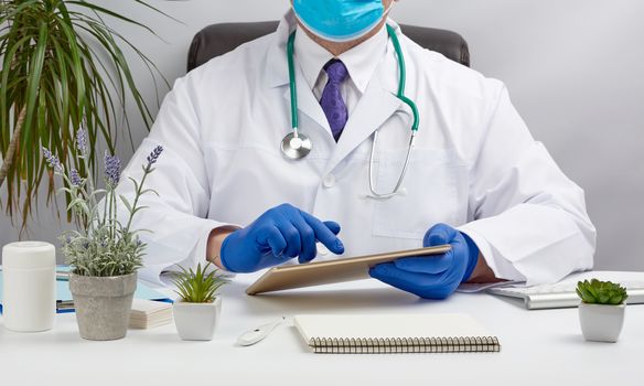 doctor in a white medical coat sits at a table and holds an electronic tablet in his hand, touches the screen with his right hand, doctor’s office, distance medicine