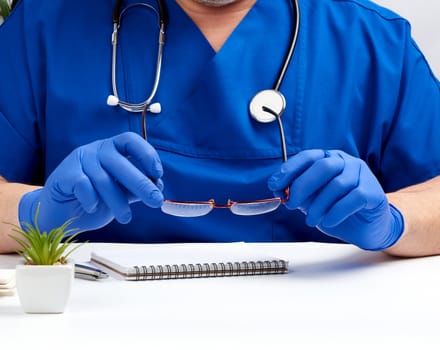doctor in blue uniform and blue latex gloves sits at a white work table in his office, concept of patient reception in the clinic, therapist's workplace