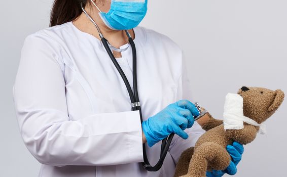 female medic holds brown teddy bear with paw bandaged in white bandage and listens to toy with stethoscope, concept of pediatrics and animal treatment