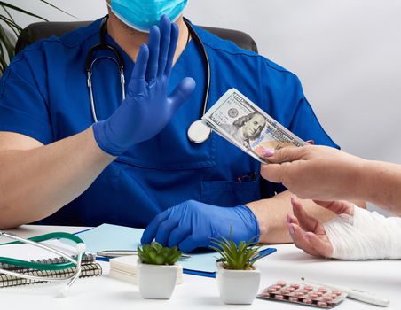 male doctor in uniform and in blue latex medical gloves sits at a table and shows with his hand a gesture of stop, hand with money stretches out from the side, concept of a bribe and expensive payment for services