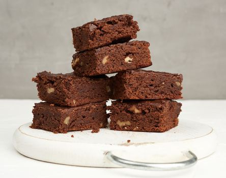 stack of square baked slices of brownie chocolate cake with walnuts on a round white wooden board