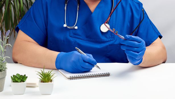 a male doctor in a blue uniform sits at a white table and writes in a paper notebook, wearing sterile gloves on his hands, concept of receiving patients in the office