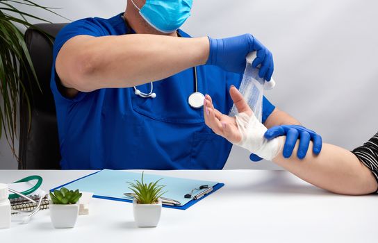male doctor sits at a table in blue uniform and latex gloves, specialist bandages his hand with a white bandage to the patient, receiving patients in the office